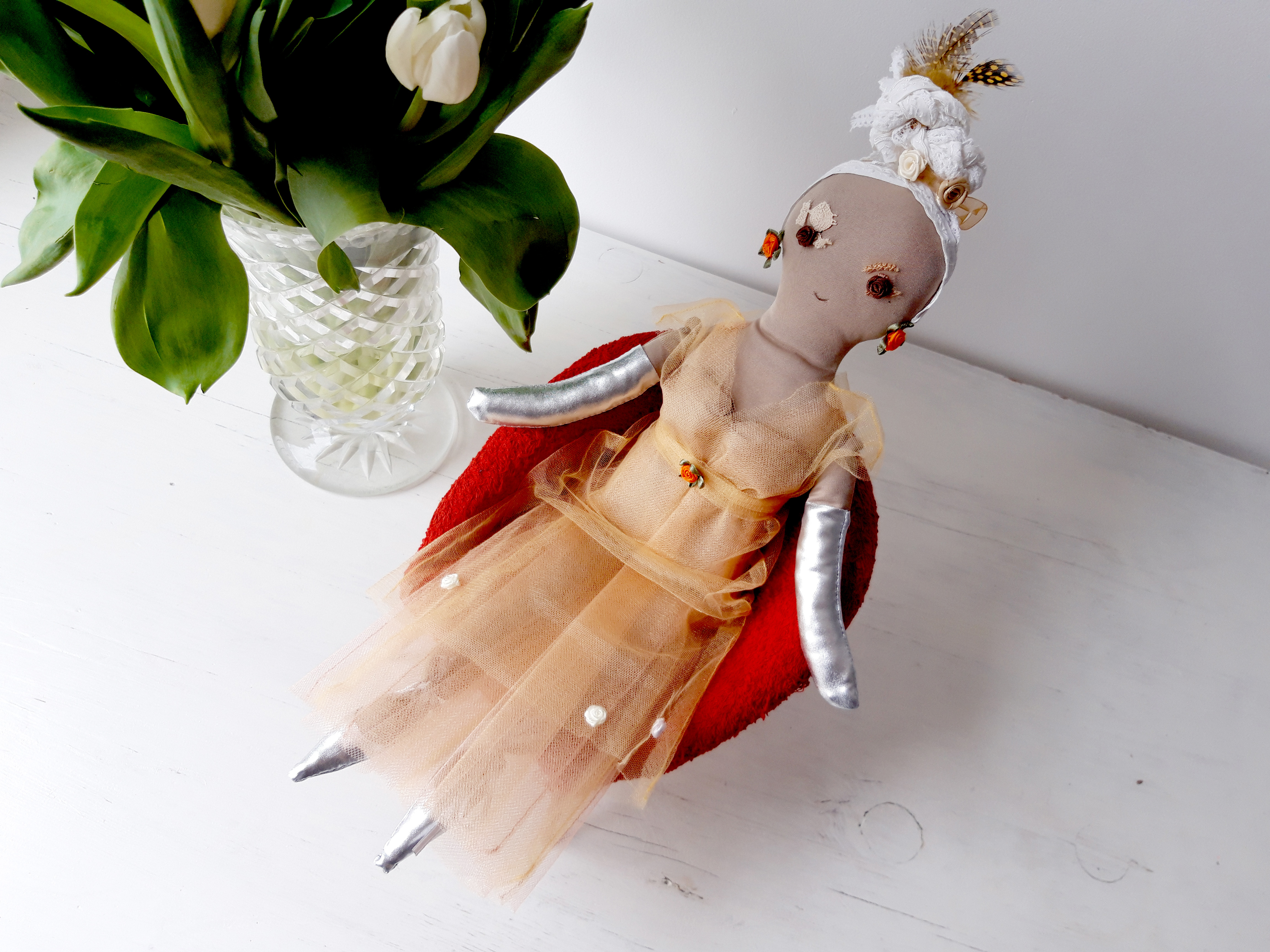 Zdjęcie przedstawia lalkę szmacianą w balowej sukni, siedzącą na foteliku dla lalek. Scena pokazana jest na białym tle. Fotelik jest koloru czerwonego. Lalka pokazana jest w pełnej postaci, en face, nieco z góry. Jest ubrana w balową suknię z tiulu w kolorze morelowym, długą do ziemi. Z lewej strony lalki, obok jej fotelika widzimy kryształowy wazon z białymi tulipanami. Białe pąki tulipanów odcinają się kontrastowo od zielonych liści.