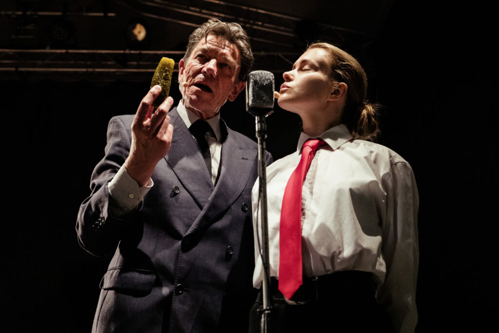 Zdjęcie. Mężczyzna w garniturze i kobieta w białej koszuli i czerwonym krawacie stoją razem na scenie, przed nimi znajduje się mikrofon. Mężczyzna trzyma w ręce kiszonego ogórka.