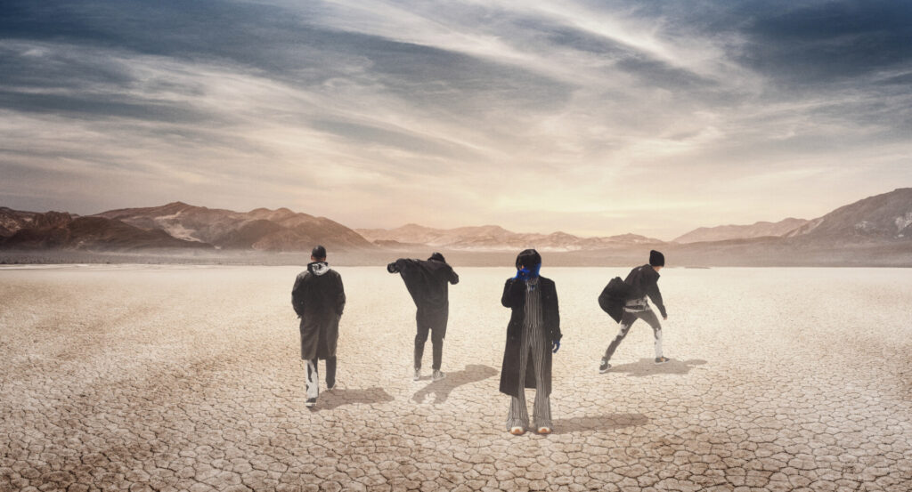 Zdjęcie. Grupa ludzi w szarych kostiumach i czarnych płaszczach znajduje się na pustyni. W oddali znajdują się skaliste wzniesienia.