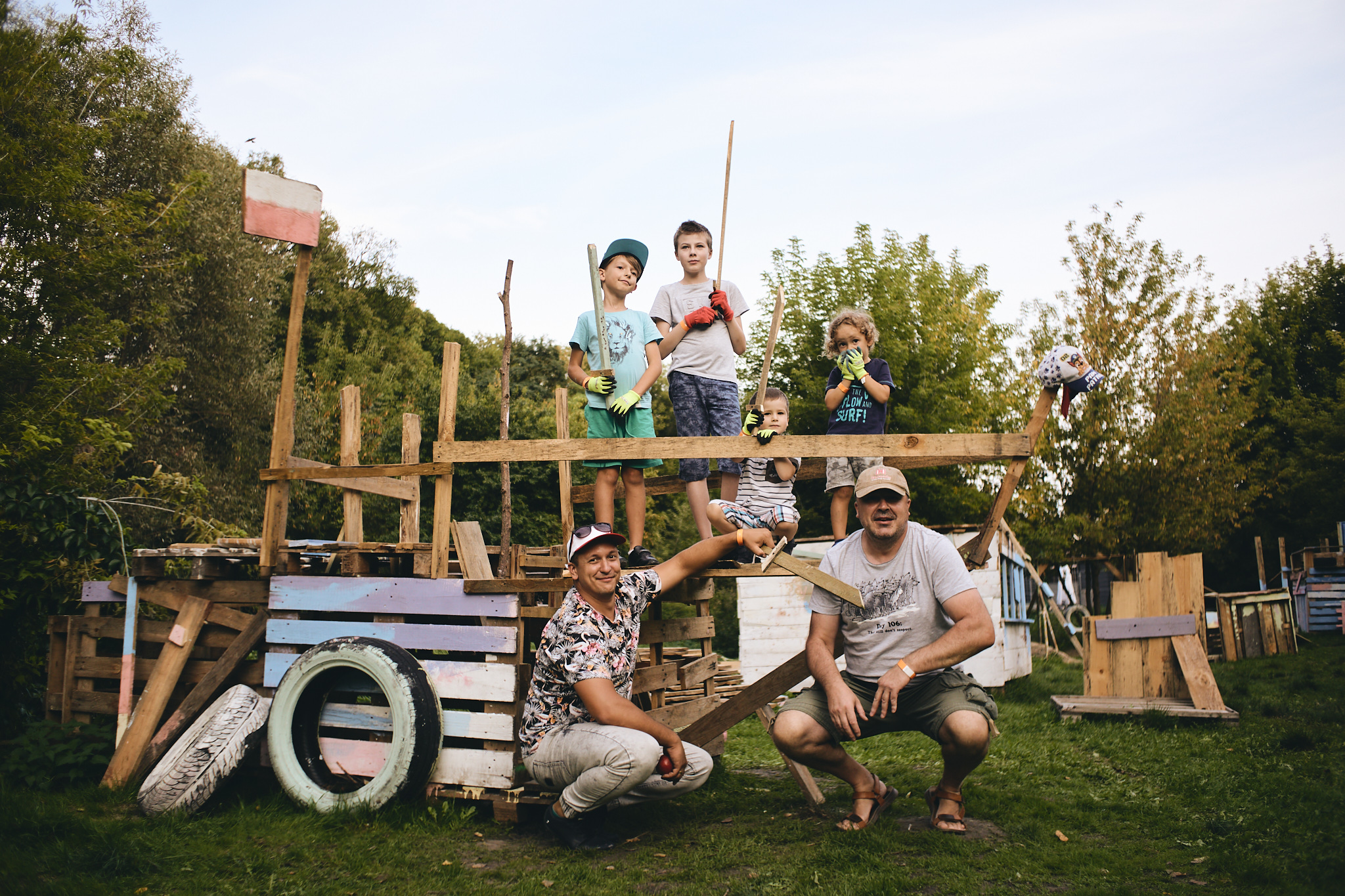 Zdjęcie z zielonego pleneru przedstawia szkielet bliżej nieokreślonej budowli z desek i palet, o którą oparte są opony. Przed budowlą przykuca dwóch mężczyzn, którzy uśmiechają się pogodnie do zdjęcia a nad nimi na ‘piętrze’ budowli jest czwórka chłopców w różnym wieku, pozują do zdjęcia. Jeden chłopiec kuca, pozostali stoją. Trzech z nich trzyma deski a jeden zasłania sobie usta rękami w roboczych rękawiczkach.
