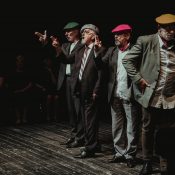 Zdjęcie ze spektaklu. Czterech mężczyzn stoi w rzędzie na scenie. Są ubrani w marynarki i kolorowe kaszkiety.