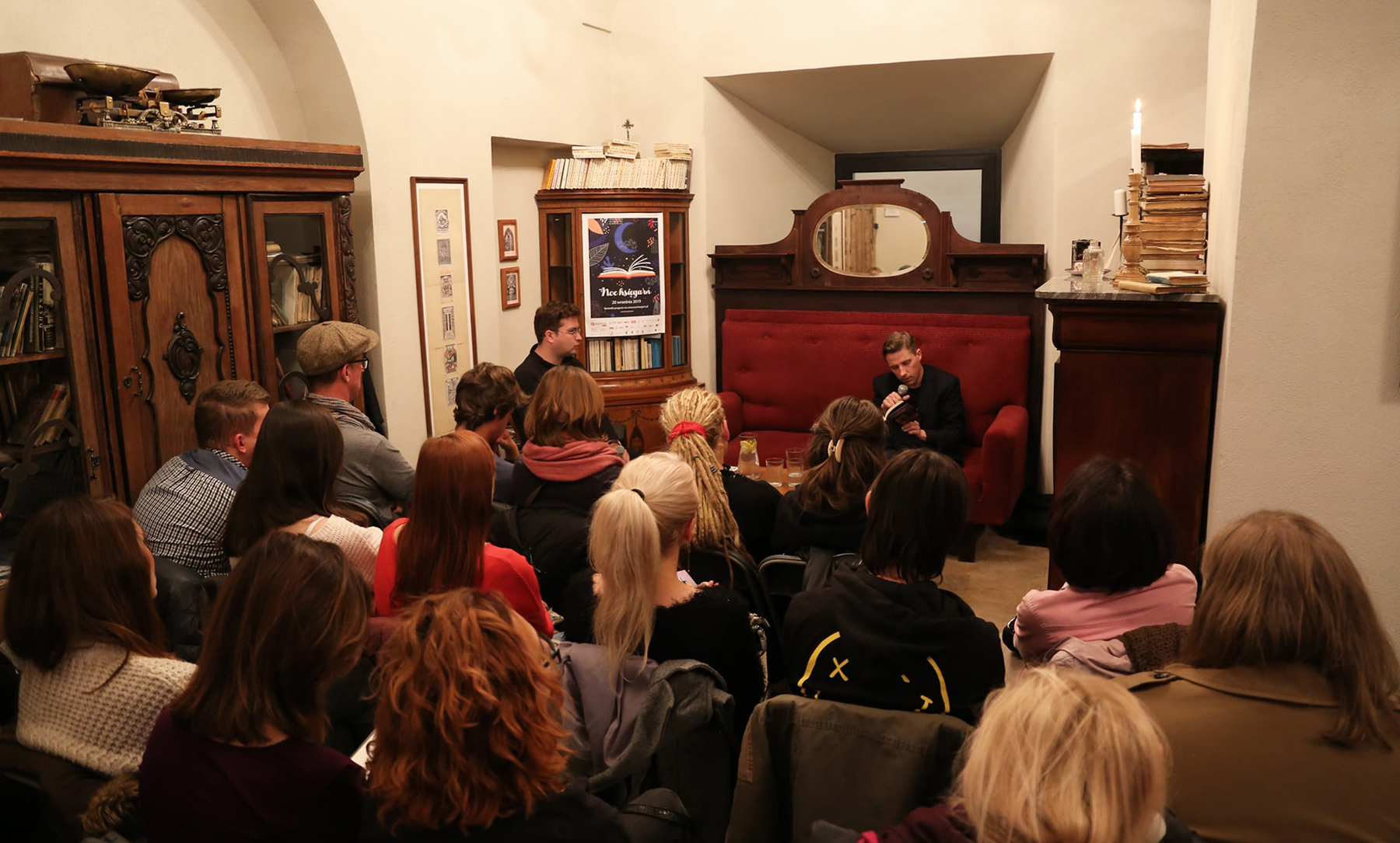 Zdjęcie przedstawia wnętrze stylizowanej na styl retro księgarni. W głębi wnętrza na stylowej obitej czerwonym materiałem kanapie siedzi mężczyzna i przez mikrofon czyta książkę, którą trzyma w dłoni. Obok niego siedzi mężczyzna prowadzący spotkanie. Naprzeciwko ich na krzesłach siedzi kilkanaście osób stanowiących widownię. Widzimy ich plecami zwróconymi do obiektywu.