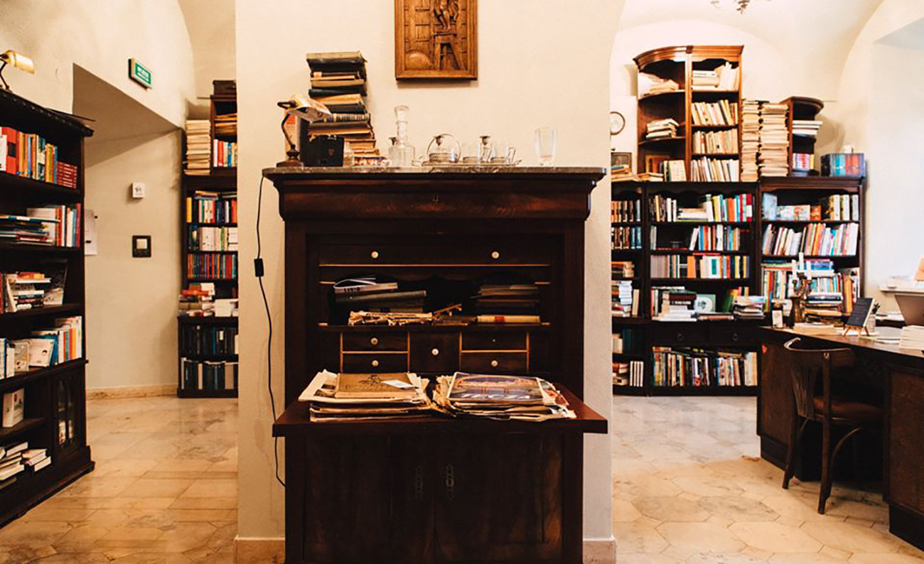 Jasne wnętrze księgarni, ciemne drewniane meble, regały wypełnione książkami, na pierwszym planie otwarty sekretarzyk z gazetami, po prawej stronie widoczne biurko.