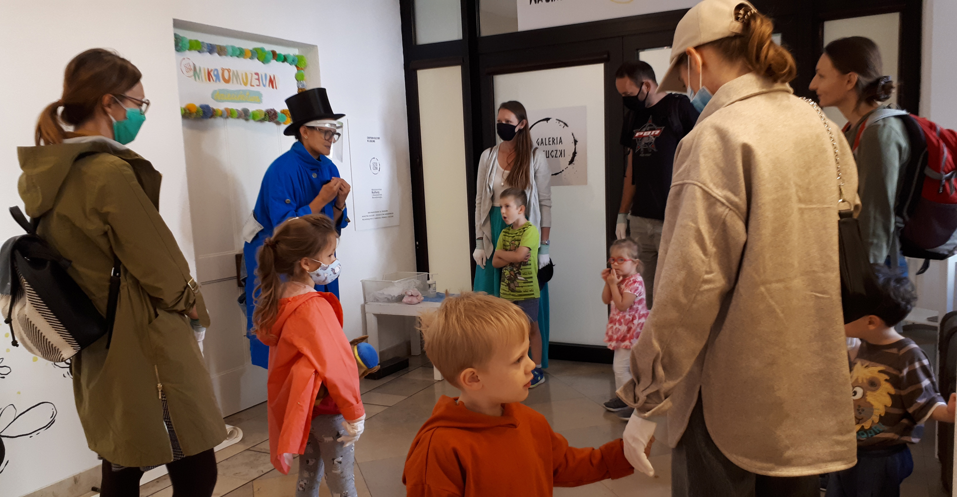 Na zdjęciu grupa dorosłych i dzieci zgromadzonych przed drzwiami z szyldem ‘MIKROMUZEUM dzieciństwa’. Przed drzwiami stoi postać w czarnym cylindrze i intensywnie niebieskim, obszernym płaszczu. Wszyscy zwróceni są ku niej a ona wydaje się coś objaśniać zgromadzonym.