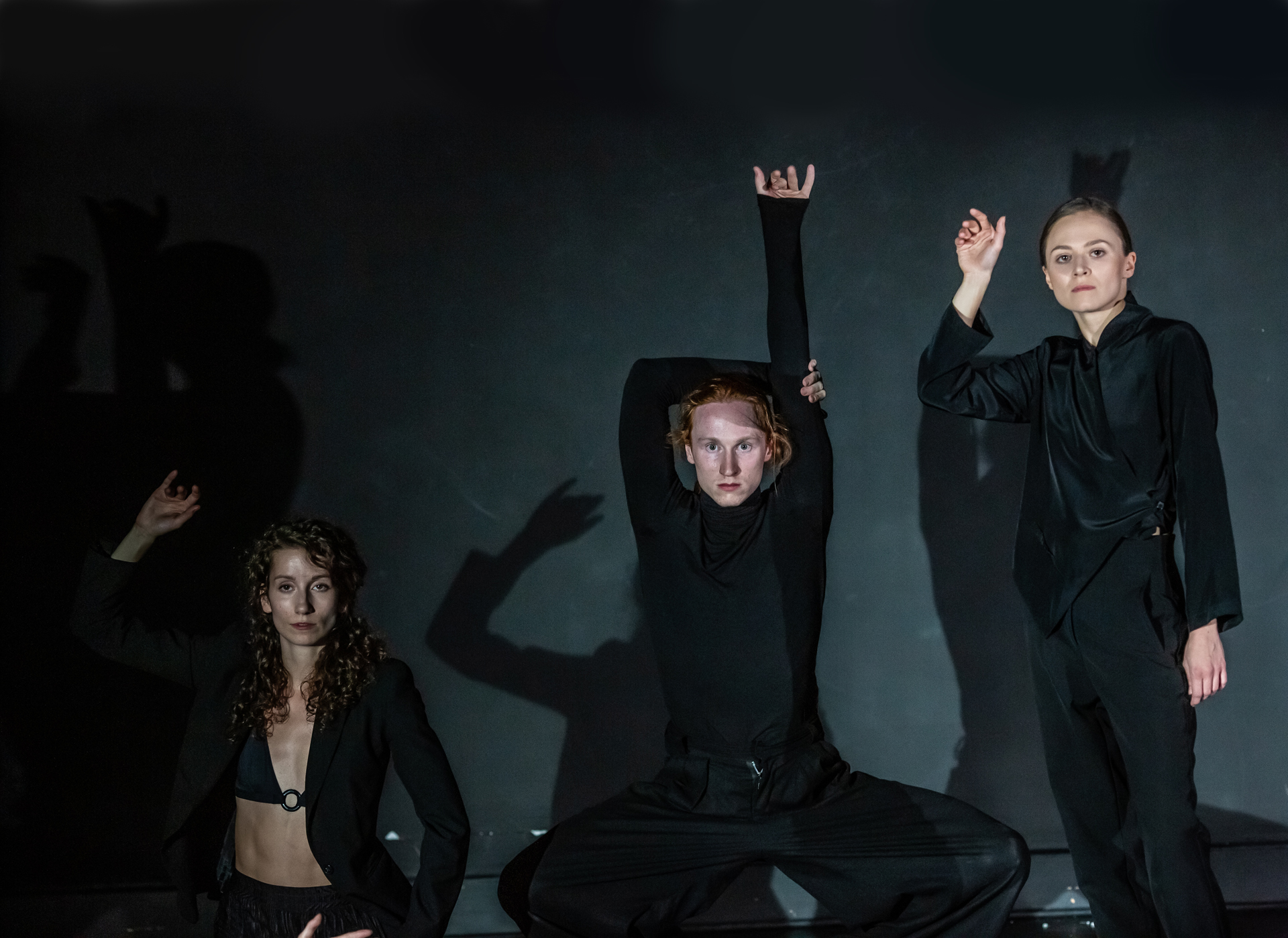 Trzy osoby ubrane na czarno w ostrym świetle, które daje odbicie ich postaci z tyłu. Kobieta z prawej stoi z podniesioną ręką, mężczyzna pośrodku kuca z kolanami na zewnątrz, uniesioną jednym ramieniem, druga ręka chwytając się za łokieć. Kobieta po lewej siedzi z uniesioną jedną ręką do góry.