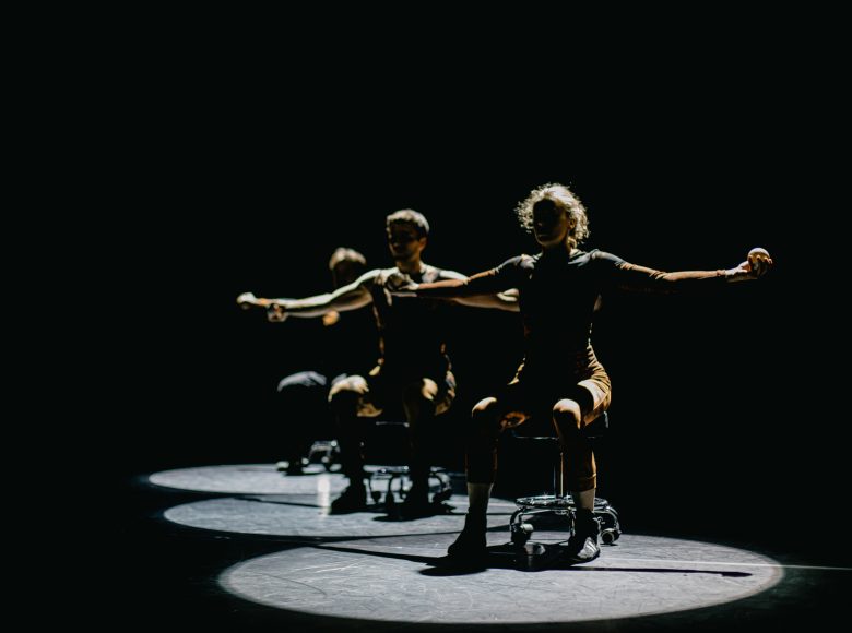 Zdjęcie przedstawia troje artystów na scenie podczas występu. Siedzą na krzesłach, ręce mają rozłożone do boku