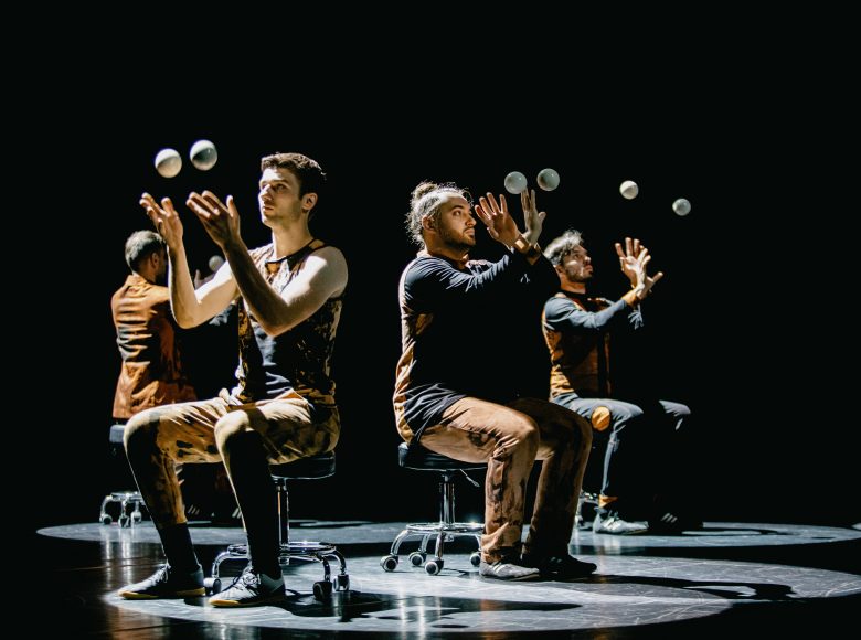 [Zdjęcie przedstawia kuglarzy żonglujących kulami na scenie. Siedzą na krzesłach, patrzą w kierunku kul