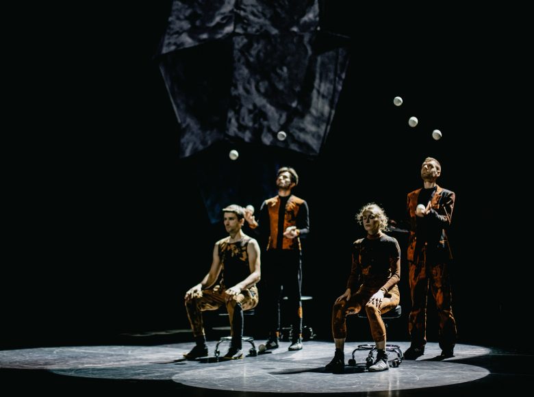 [Zdjęcie przedstawia kuglarzy żonglujących kulami na scenie. Siedzą na krzesłach, patrzą w kierunku kul