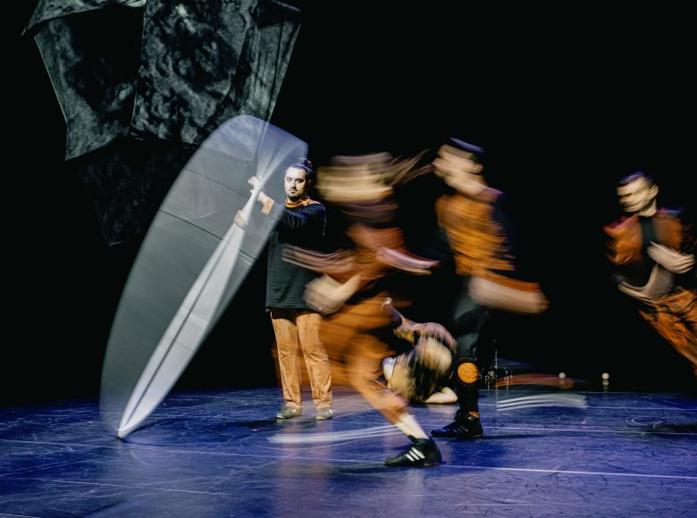 Na scenie występ kuglarzy. Za nimi czarne tło.