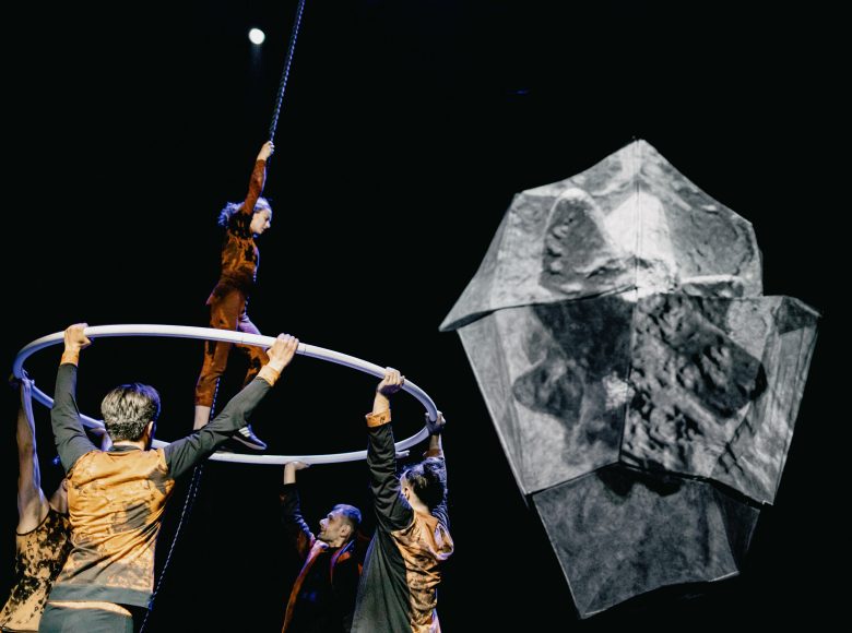 Zdjęcie przedstawia kuglarzy podczas występu na scenie. Mężczyźni trzymają wielkie koło, po którym chodzi akrobatka