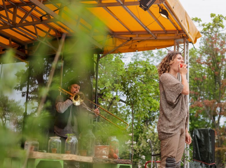 Zdjęcie aktorzy na scenie. Kobieta stoi z przodu i gryzie jabłko. Mężczyzna za nią gra na puzonie.