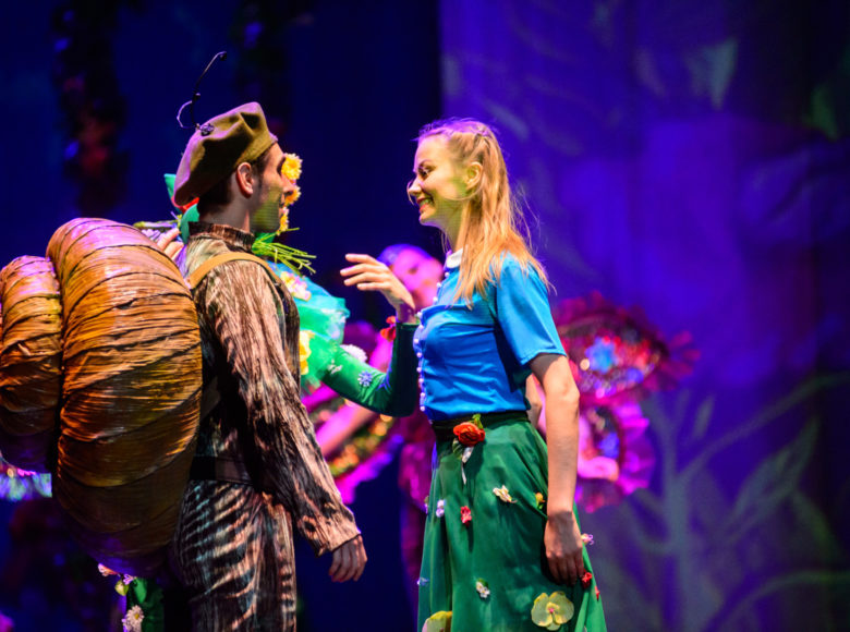 [Zdjęcie przedstawia scenę teatralną. Aktor po lewej stronie ubrany jest w kostium ślimaka. Po lewej stronie stoi uśmiechająca się do niego aktorska w niebieskiej bluzce z giziczkami i spódnicy z przyczepionymi do niej sztucznymi kwiatami]