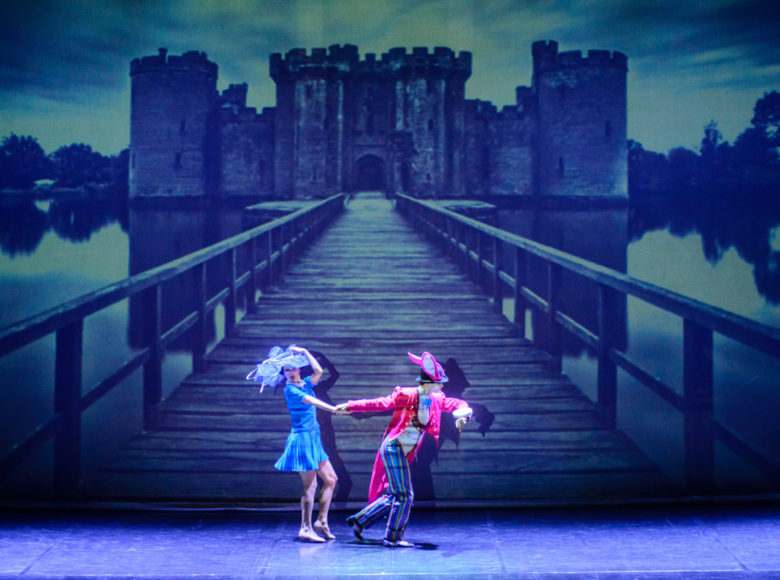 Zdjęcie przedstawia parę aktorów na scenie. Kobieta ma niebieską sukienkę i kapelusz. Mężczyzna ubrany jest w strój królika i ciągnie kobietę za rękę. W tle znajduje się zdjęcie zamku i prowadzącego do niego mostu