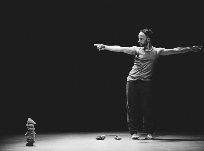 Czarno-biała fotografia. Scena. Po prawej stronie stoi mężczyzna. Ma bose stopy, czarne długie spodnie i szarą koszulkę na ramiączkach. Stoi przodem. Ręce ma uniesione na boki. Patrzy w lewą stronę. Po lewej stronie sceny ułożona jest mała wieża z kilku kamieni.