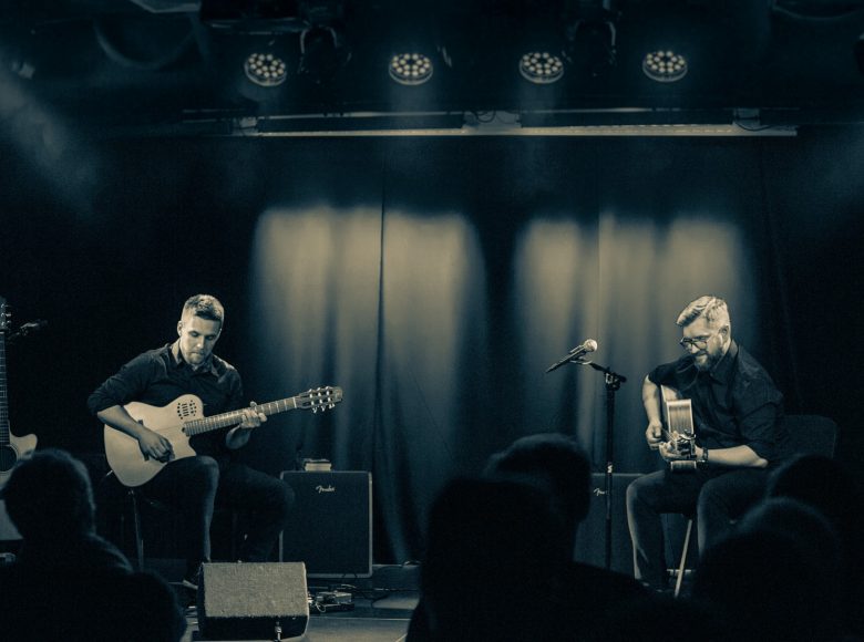 Na zdjęciu w odcieniach szarości widzimy dwoje mężczyzn z gitarami siedzących na krzesłach na scenie