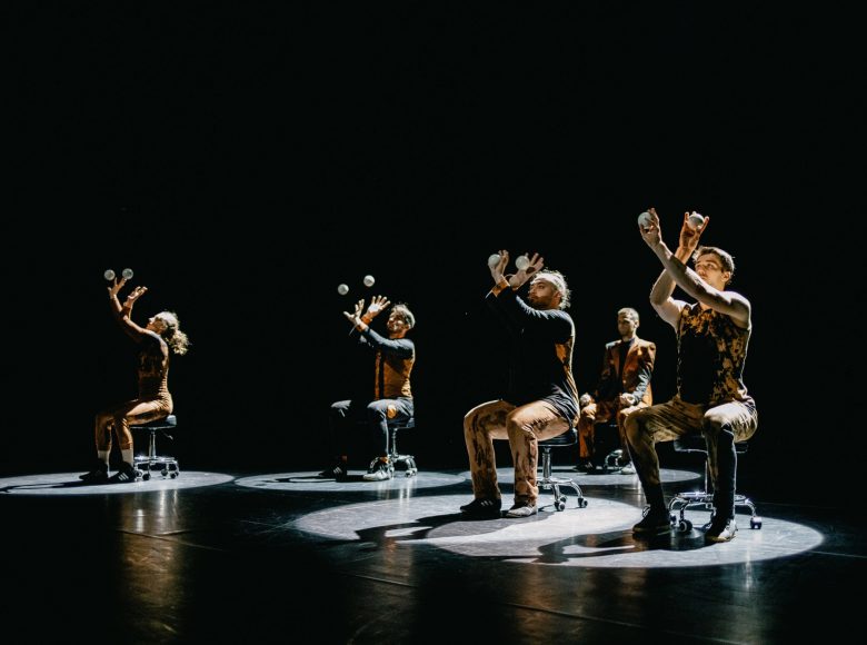 [Zdjęcie przedstawia kuglarzy żonglujących kulami na scenie. Siedzą na krzesłach, patrzą w kierunku kul