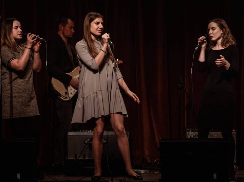 [Zdjęcie przedstawia cztery osoby na scenie. Mężczyzna w tle gra na gitarze. Trzy młode kobiety na przodzie sceny śpiewają.]