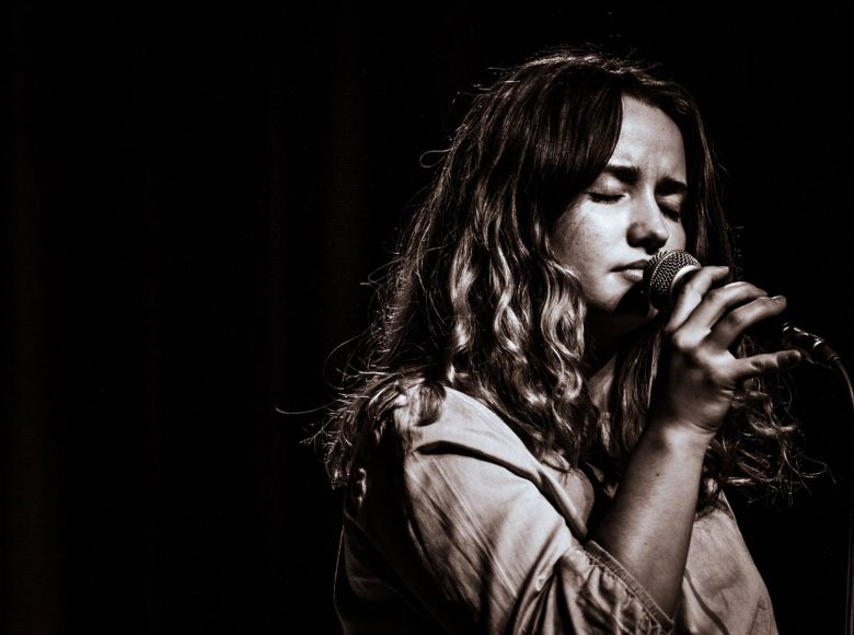 [Zdjęcie w sepii. Przedstawia młodą kobietę, która śpiewa na scenie. W ręku trzyma mikrofon. Ma zamknięte oczy]