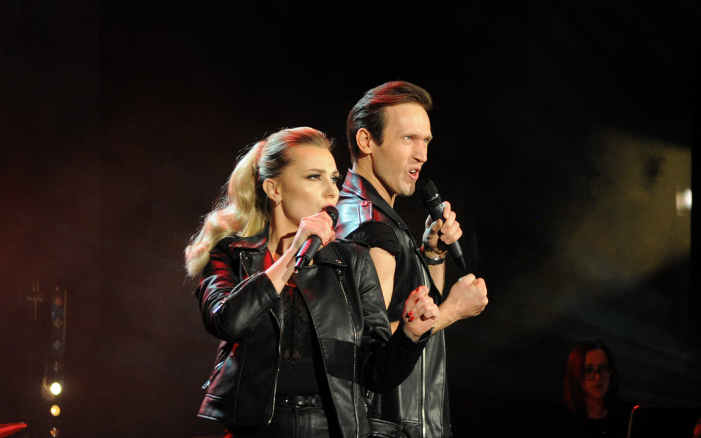 [Zdjęcie przedstawia kobietę i mężczyznę śpiewających na scenie. Są ubrani w czarne skórzane kurtki]