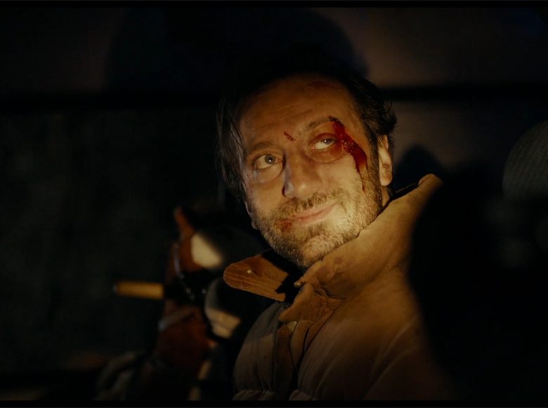 [Kadr z filmu. Noc. Zdjęcie przedstawia mężczyznę z podbitym okiem. Po policzku spływa mu strużka krwi]