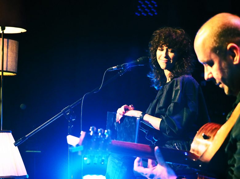 Na scenie wokalistka i gitarzysta. Oświetleni są punktowym światłem, za nimi czarne tło.