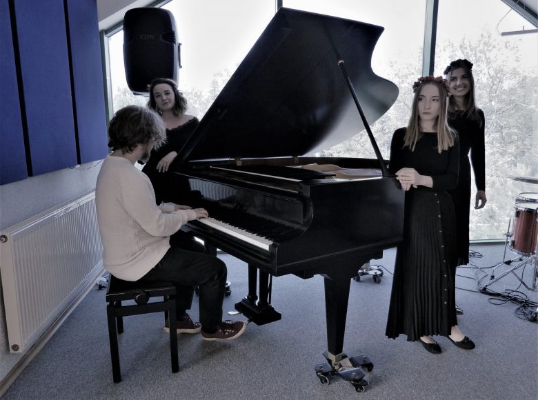 Trzy młode dziewczyny oparte o fortepian uśmiechają się szczerze. Ubrane są w ciemne sukienki. Dwie z nich o długich włosach na głowach mają nałożone wianki