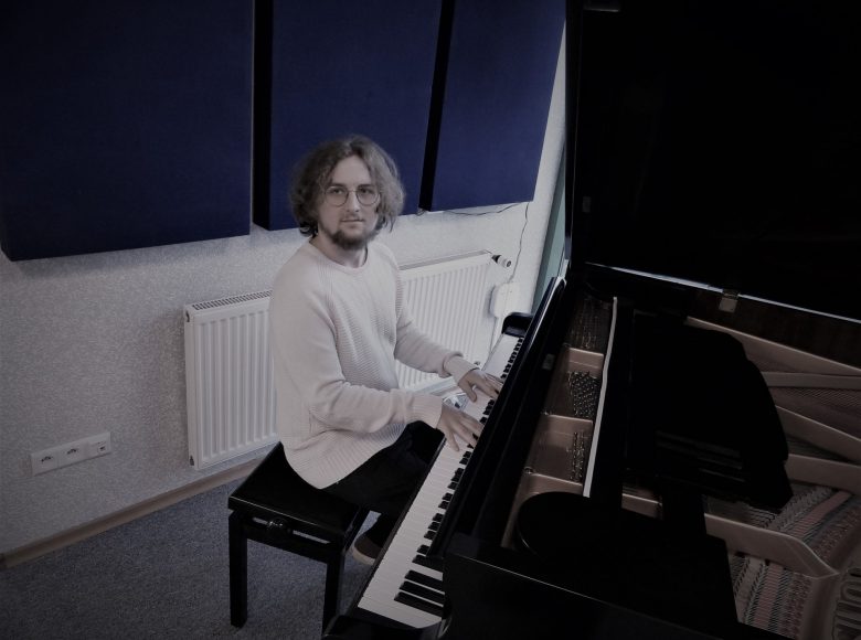 Przy czarnym fortepianie siedzi młody mężczyzna w okularach. Ma długie blond włosy. Ubrany jest w biały sweter i ciemne spodnie. W tle jest biała ściana z niebieskimi kwadratowymi panelami i szara podłoga