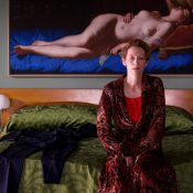 Kobieta ubrana w ciemnoczerwony szlafrok siedzi na łóżku. Łóżko przykryte zieloną narzutą, po bokach dwie lampki nocne. Nad łóżkiem obraz nagiej kobiety w stylu renesansowym.