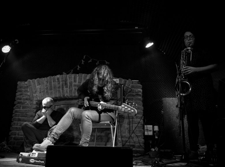 Czarno-białe zdjęcie przedstawia muzyków na scenie. Meżczyzna po prawej siedzi w okularach na podłodze. Mężczyzna w środku siedzi na krzesełku i gra na gitarze. Kobieta po lewej stronie gra na saksofonie]