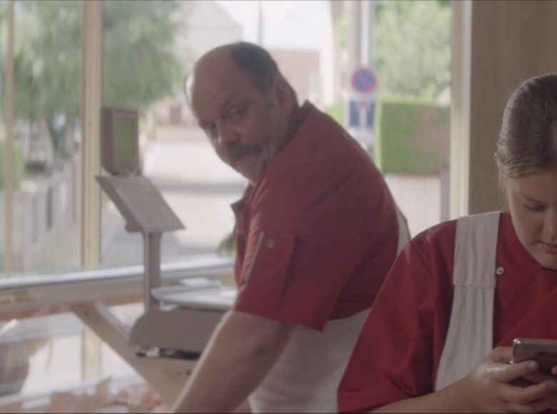 [Kadr z filmu. Wnętrze sklepu. Przy ladzie z wagą stoi mężczyzna, który spogląda na dziewczynę patrzącą się w ekran telefonu]