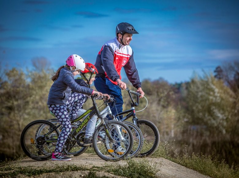 [Dzień. Zdjęcie przedstawia trzy osoby - dorosłego mężczyznę i dzieci jadące na rowerach po ścieżce wśród drzew]