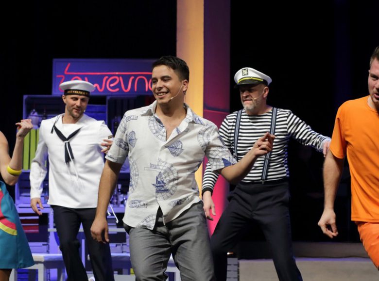 Na scenie grupa tańczących osób, jedna kobieta i kilku mężczyzn. Trzech mężczyzn ubranych jest w stroje marynarzy.