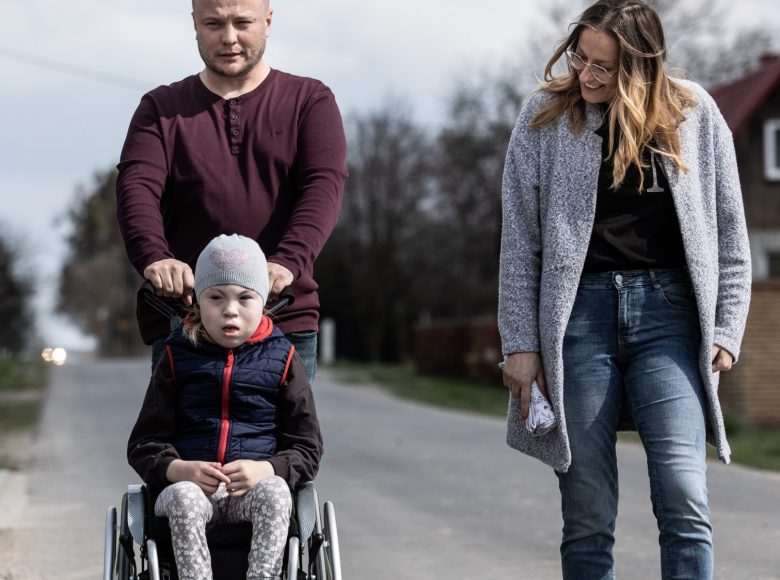 [Zdjęcie przedstawia rodzinę. Mężczyzna pcha wózek z dziewczynką. W ich kierunku patrzy uśmiechnięta kobieta]