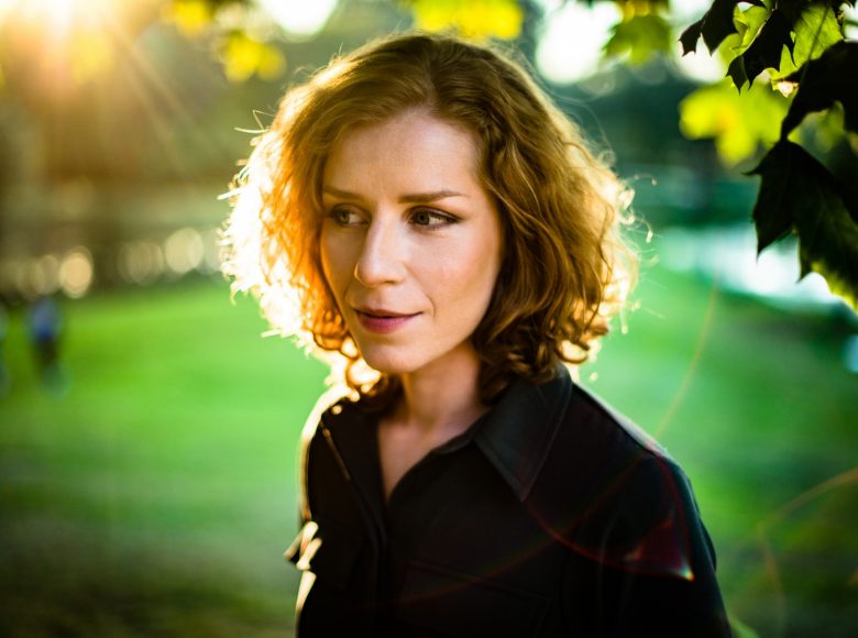 Fotografia wokalistki Aliny Orlove. Młoda kobieta ma falowane, ciemnoblond włosy do ramion, uśmiecha się, kieruje spojrzenie w lewą stronę kadru. Widać jej głowę i ramiona, ma ciemną bluzkę i wyraźny makijaż. Światło pada na jej twarz, tak ja promienie słoneczne.