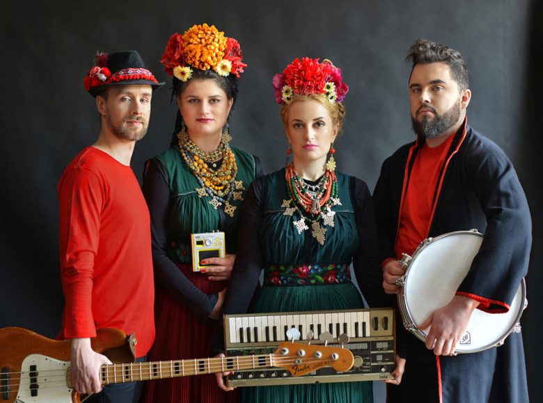 Zdjęcie. Czterej członkowie zespołu Dagadana ubrani są w ludowe stroje i trzymają w rękach instrumenty muzyczne. Kobiety na środku mają wianki ze świeżych kwiatów