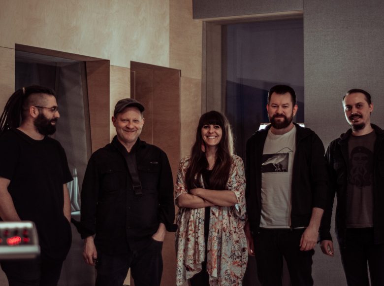 [w przestrzeni studia nagraniowego stoi pięcioro osób: czworo mężczyzn a w środku kobieta]