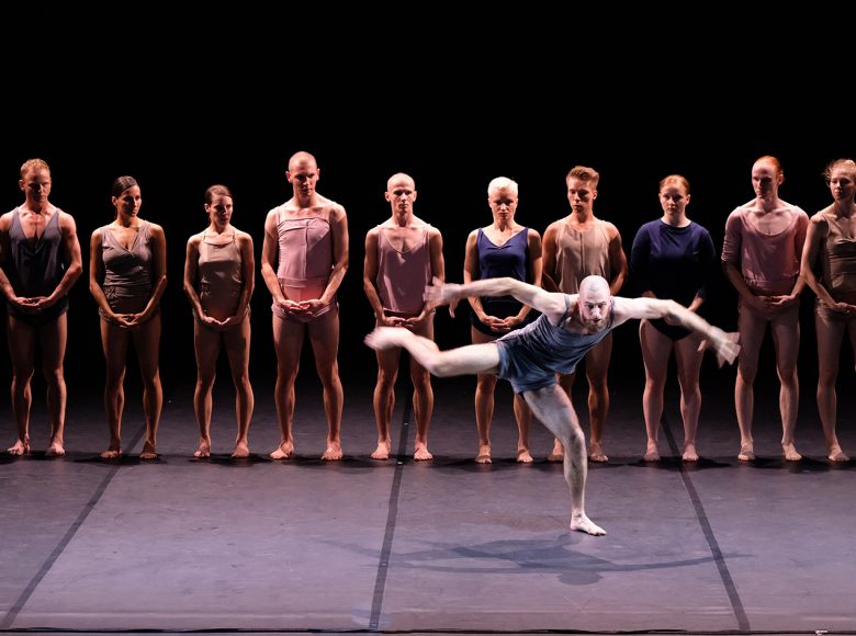 Zdjęcie. Tancerze na scenie stoją w równym rzędzie. Przed nimi inny tancerz wykonuje układ taneczny. Pochyla się. Stoi na jednej nodze. Drugą nogę ma wyprostowaną do boku