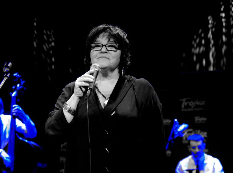 Zdjęcie z koncertu. Artystka z krótkimi włosami i w okularach korekcyjnych stoi przy mikrofonie. W tle mężczyźni oświetleni niebieskim światłem grają na instrumentach