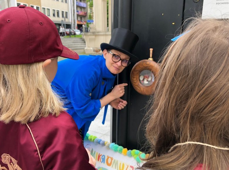 Zdjęcie. Kobieta w niebieskim płaszczu, okularach i meloniku pokazuje coś dziewczynkom w trakcie warsztatu.