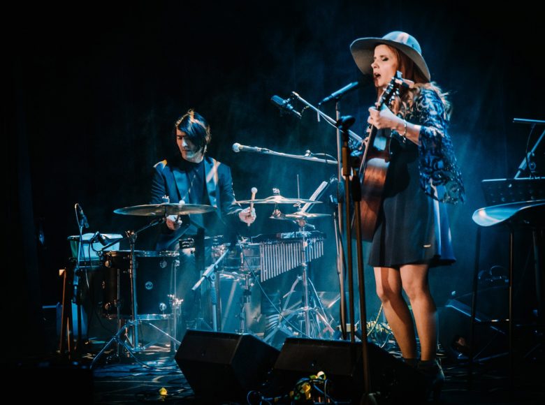 Dorota Osińska śpiewa na niebiesko oświetlonej i spowitej dymem scenie. Ma zamknięte oczy, stoi pryz mikrofonie i gra na gitarze. Ma długie rude włosy, krótką sukienke i kapelusz z dużym rondem. W tle, w prawej części fotografii widać perkusistę, który gra na instrumencie.