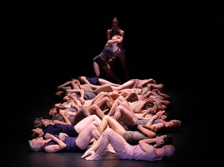 Zdjęcie ze spektaklu tanecznego. Tancerze leżą w dwóch rzędach na scenie. Mają podkulone nogi, a ich ręce przylegają do klatki piersiowej
