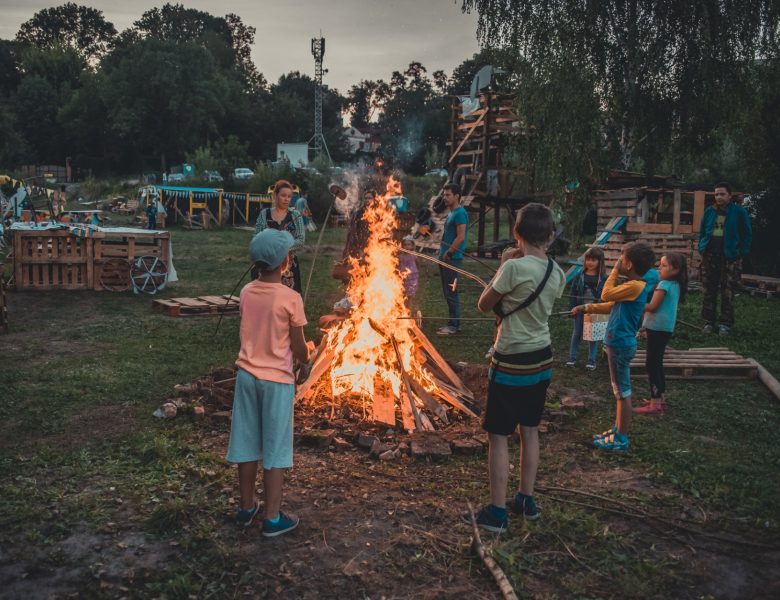Wieczór. Zdjęcie przedstawia grupę dorosłych i dzieci, którzy pieką kiełbaskę nad ogniskiem. Wokół nich znajdują się drewniane konstrukcje i drzewa.