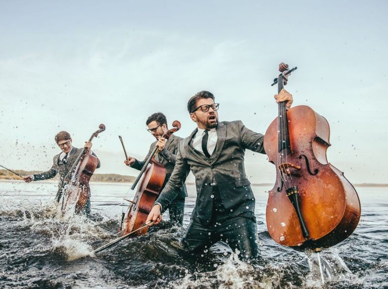 Trzech mężczyzn w garniturach. Stoją po kolana w wodzie. W rękach trzymają wiolonczele. Zdjęcie jest dynamiczne.