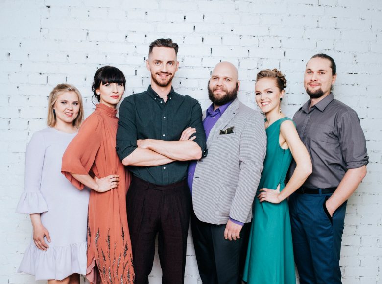 Wokaliści zespołu Estonian Voices pozują na tle ściany z cegieł pomalowanej na biało. Trzy kobiety i trzech mężczyzn uśmiechają się i noszą kolorowe ubrania