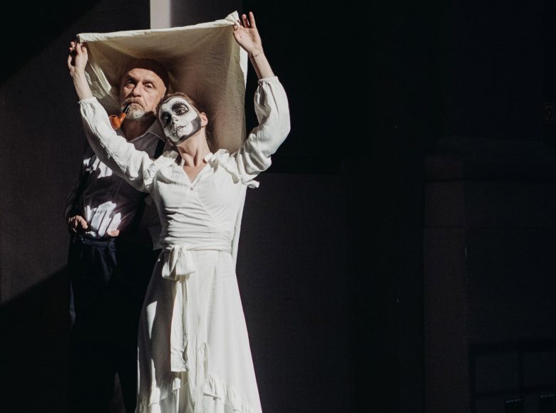 Zdjęcie. Scena ze spektaklu "Margules. Nieobecność". Kobieta w białej sukience ma sceniczny makijaż przedstawiający trupia czaszkę. Obok niej stoi mężczyzna z drewnianą fajka w ustach. Kobieta osłania siebie i mężczyznę biała chustą. Scena jest ciemna, w rogu widać fragment drewnianych drzwi.
