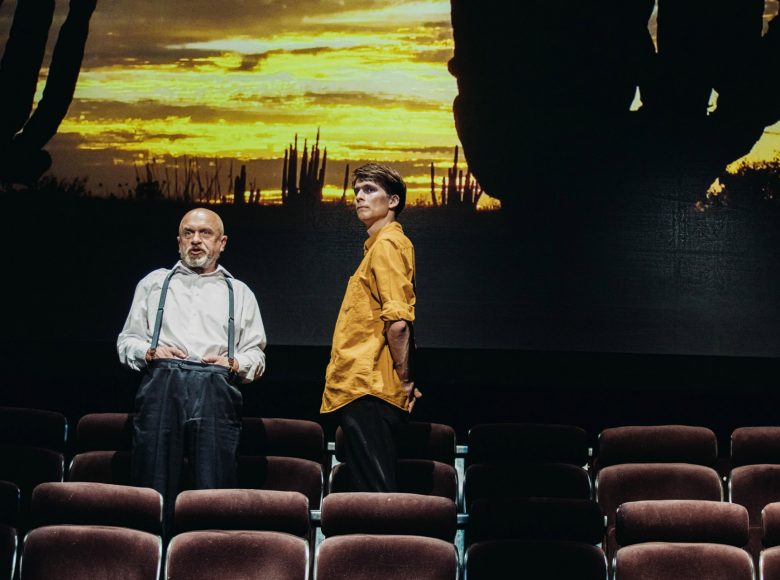 Zdjęcie. Scena ze spektaklu "Margules. Nieobecność". Dwóch mężczyzn stoi na tle zachodzącego słońca. Starszy z nich jest ubrany w białą koszulę i spodnie na szelkach. Ręce trzyma za paskiem spodni. Młodszy mężczyzna stoi bokiem. Jest ubrany w żółta koszulę i ciemne spodnie. Jego twarz jest pomalowana białym pudrem, ma podkreślone kości policzkowe i pomalowane na ciemno oczy. Mężczyźni stoją między rzędami siedzeń.