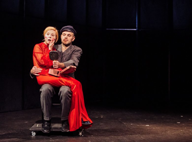 Scena ze spektaklu ":Biesy". Aktor siedzi na krześle, w ręku trzyma lusterko. Na jego kolanach siedzi kobieta w czerwonej sukni.