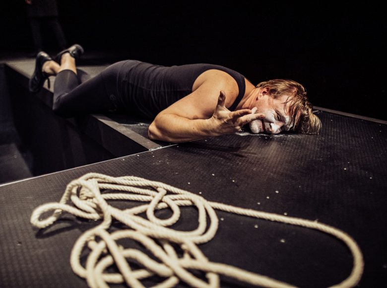 Scena ze spektaklu "Biesy". Aktor z pomalowaną na biało twarzą leży na scenie. Przed nim znajduje się leżący w nieładzie sznur.