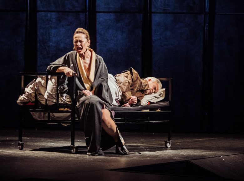 Scena ze spektaklu "Biesy". Aktor leży na szpitalnym łóżku. Na brzegu łózka siedzi odwrócona do niego plecami kobieta i krzyczy.