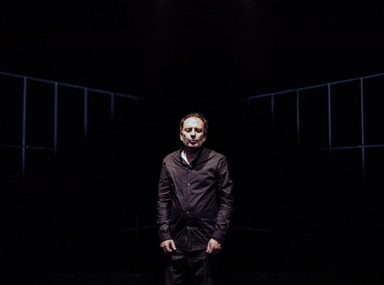 Scena ze spektaklu biesy. Na środku sceny stoi aktor. Ma zamknięte oczy, jest ubrany na czarno. Na jego twarz pada światło. Aktor patrzy w dół.